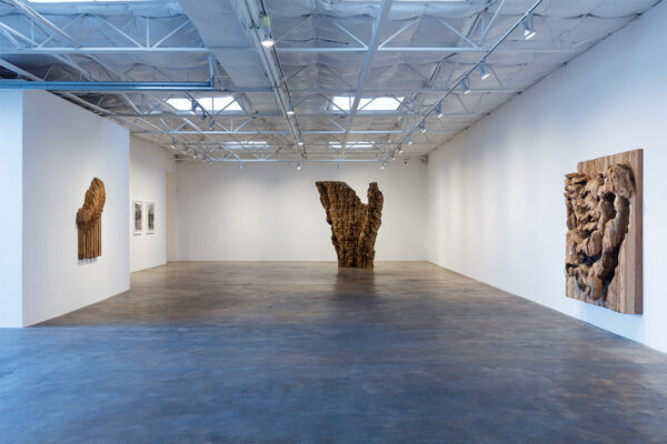 An installation view of work by Ursula von Rydingsvard. A large freestanding wooden sculpture sits at the back of the gallery and two smaller relief sculptures hang on the gallery walls.