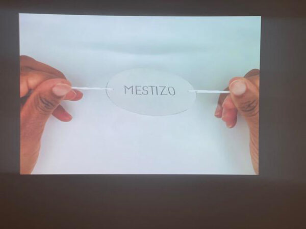 Video still of the artist holding two threads on each side of a paper that says mestizo