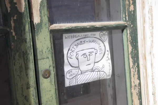 A green door with a hand-drawn paper sign with text that reads, "WebbGallery.com Fort Davis-Waxahachie." There is a line drawing of a man wearing a cowboy hat. 