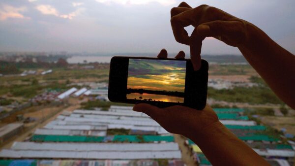 A still image from a digital video by Sindhu Thirumalaisamy. The image shows a blurred background of what appears to be rows of buildings. In the foreground, a person holds a cell phone that displays an image of a colorful sunset.