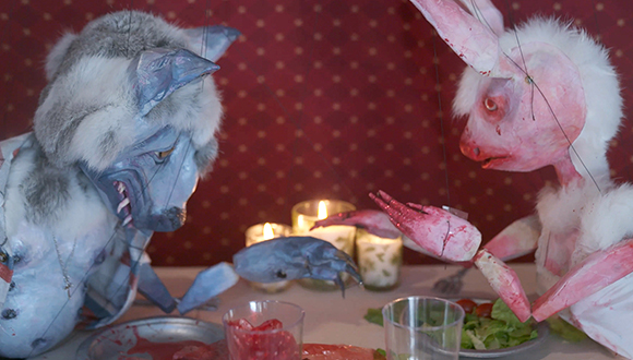A still image from the video "Bad Bunny," by Sarah Fox. The image shows two puppets of anthropomorphized animals eating dinner at a restaurant.