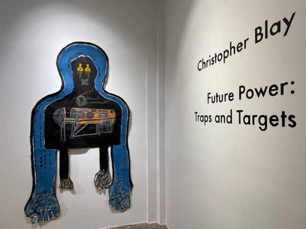 A photograph of a work of art by Christopher Blay. The mixed media work hangs on the gallery wall. It depicts an abstracted human form from the chest upward with hands dangling below. To the right of the work is text that reads, "Christopher Blay Future Power: Traps and Targets."