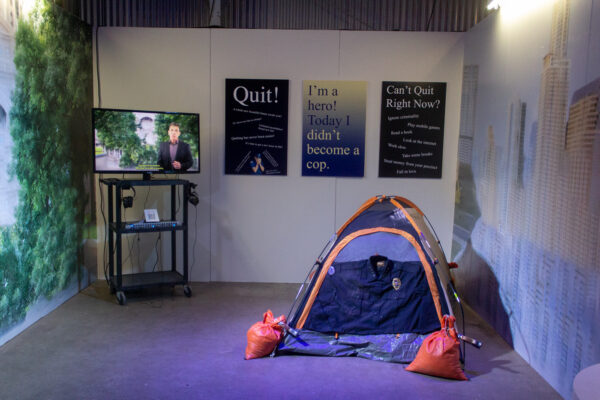 An installation view of a sculptural collaboration between Andie Flores and Sam Levine. A Tv on a stand sits in the corner, three didactic prints hang on the wall, and a tent with sandbags is set up on the floor.