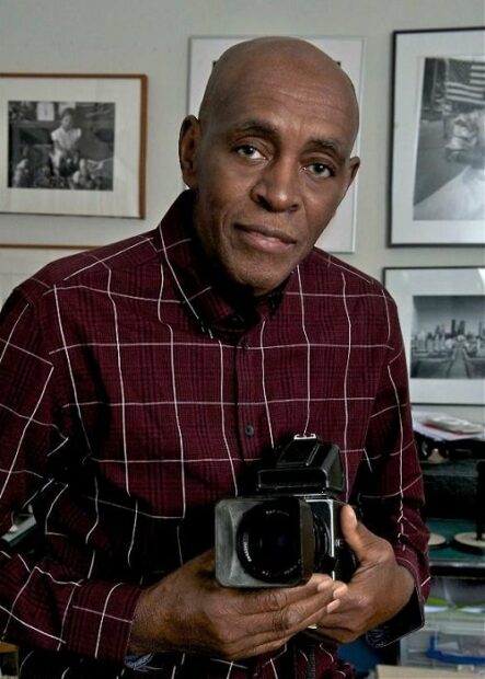 A portrait of artist Earlie Hudnall Jr. The artist wears a plaid long sleeve shirt and holds a camera while he looks straight at the viewer.
