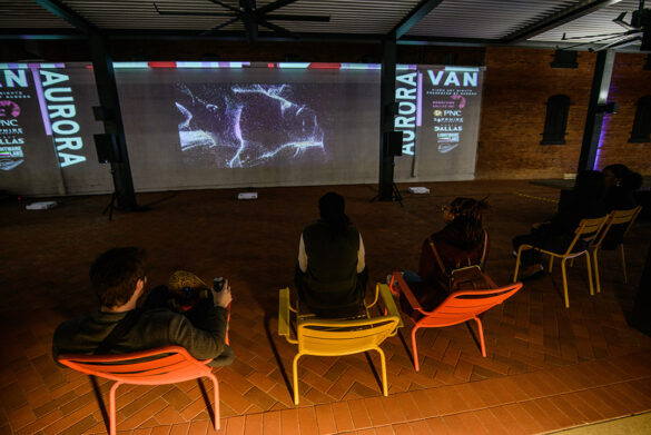A photograph of a group of people seated in colorful chairs and watching a projection of an abstract video work.