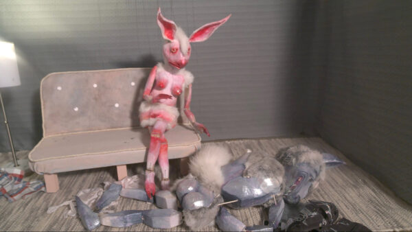 A still image from a video work by Sarah Fox. The image depicts two anthropomorphized animal puppets, a partially nude female rabbit sitting on a couch as she looks at a nude male wolf lying on the ground face down.