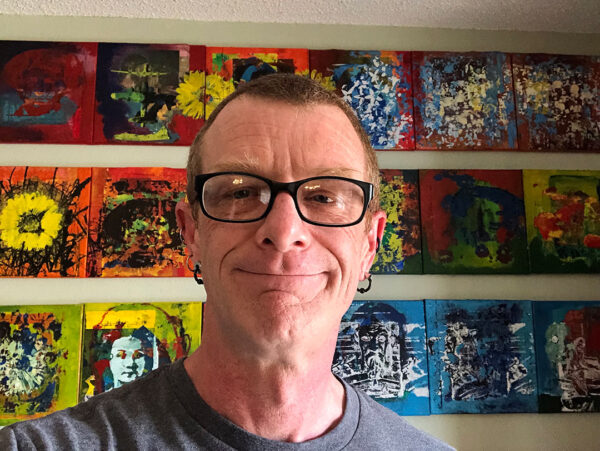 A photograph of Dr. Christian Conrad. He wears black-rimmed glasses and a looks directly into the camera with a slight smile. There are three rows of colorful prints on the wall behind him. 