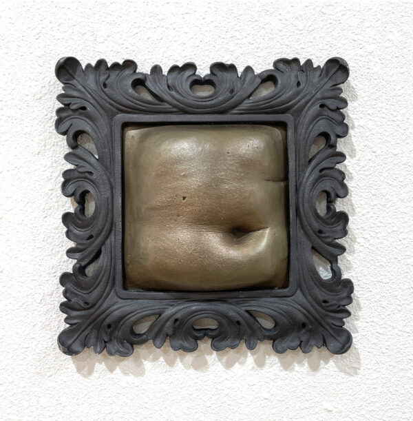 A photograph of a relief sculpture by Erin Cunningham. A bronze cast frame hangs on a white wall. The frame is filled by a plaster cast of a human stomach which has been painted a lighter bronze color.