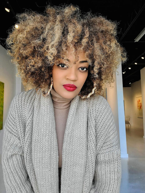 A headshot of Daisha Board, owner and director of Daisha Board Gallery. Board wears a light gray sweater cardigan and large hoop earrings. She looks directly in the camera with a slight smile.