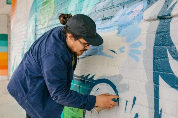 A photograph of artist David Maldonado painting a mural with a can of spray paint.