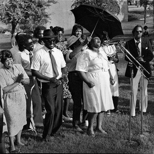 A black and white photograph by Earlie Hudnall Jr. The image depicts a small group of people standing outdoors and singing in front of a microphone.