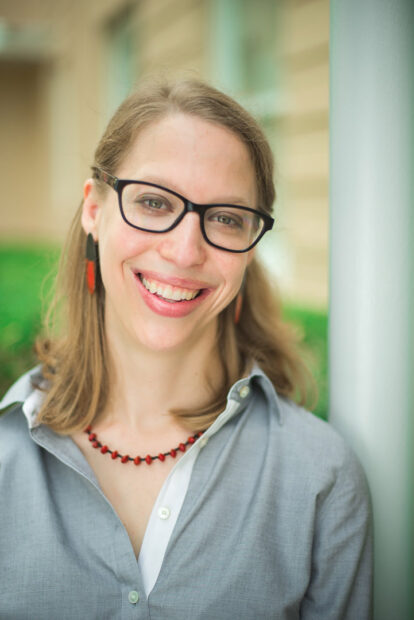 A headshot of Kathryn Hall. She wears a button-up collared shirt, a red necklace and red earrings, and simple black glasses. She looks at the camera and smiles widely.