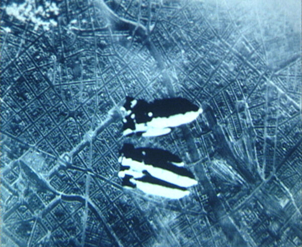 A screen shot from a film by Harun Farocki. The black and white image is taken from a plane and appears to show two bombs that are being dropped.