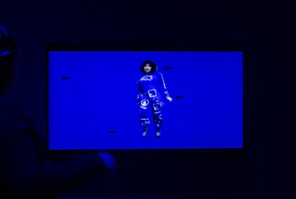 A photograph of a television screen. A woman in a blue bodysuit stands amid a blue background, mid-movement