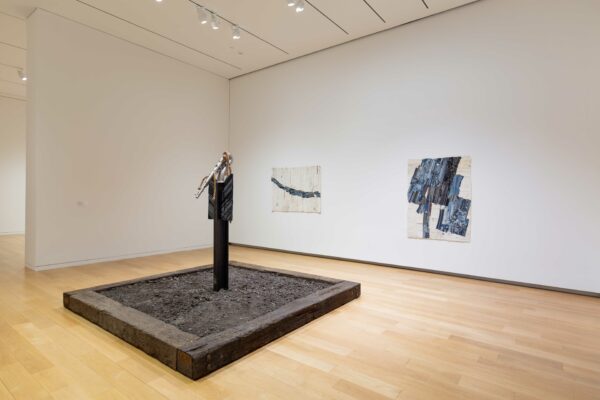 Installation view of two dimensional works on a wall with a sculpture of a saxophone in the foreground