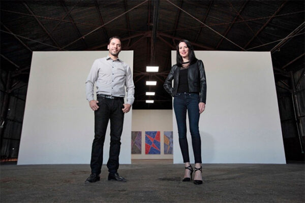 A photograph of Jason and Nancy Koen standing inside a gallery space. Behind them are white walls and in the background three colorful abstract works of art are visible.
