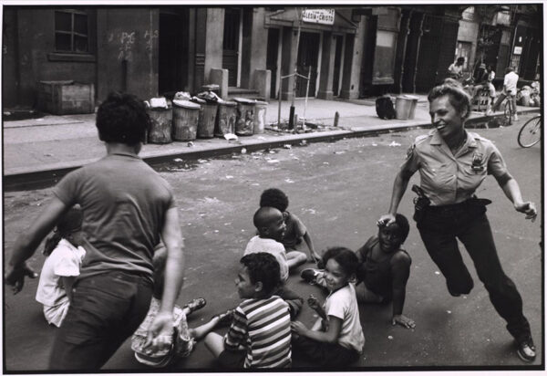 A black and white photograph by Leonard Freed of a policewoman playing with a group of young children. Most of the children are seated and the policewoman smiles as she chases one child around the seated children.