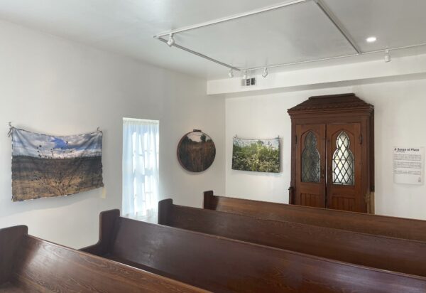 Vista de un cuarto con paredes blancas, una entrada de dos puertas con ventanas góticas y bancos de iglesia de madera. De las paredes cuelgan paisajes.