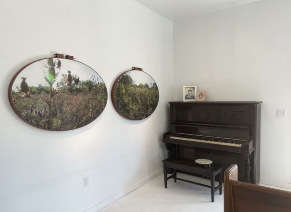 En las paredes de la esquina de un cuarto blanco está instalado un piano vertical de madera oscura sobre el cual hay un par de retratos enmarcados. Sobre la pared perpendicular al piano hay dos paisajes ovalados.