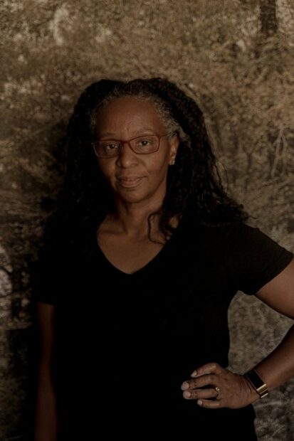 A portrait of Letitia Huckaby. The artist wears a black short sleeve shirt and has one hand on her hip while she looks straight into the camera. 