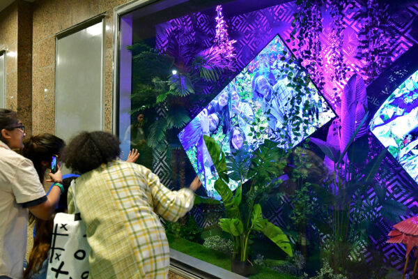 A photograph of three people interacting with an installation by Lina Dib. The viewers stand in front of the installation and see their images played back on a screen in front of them.