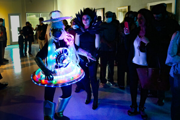 A photograph of a group of people in a darkened gallery space. The main subject of the image is a person dressed in a whimsical light-up costume which includes a headpiece that resembles a ringed planet, a skirt that resembles a UFO with lights, and puffy silver boots. 