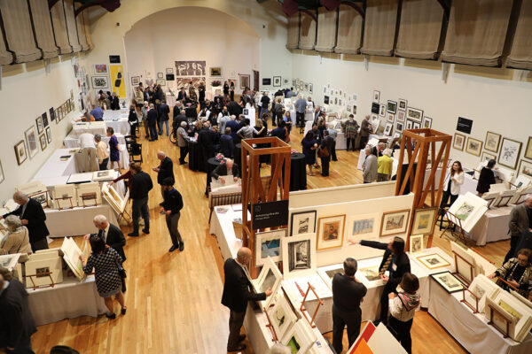 A photograph of a large room with several booths set-up selling prints. Crowds of people move around the space looking at prints and talking.