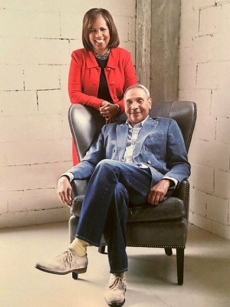A photograph of Melanie Lawson and John Guess Jr. Mr. Guess sits in a chair with his legs crossed and smiles as he looks into the camera. Ms. Lawson stands behind the chair and has a wide smile as she looks directly into the camera.