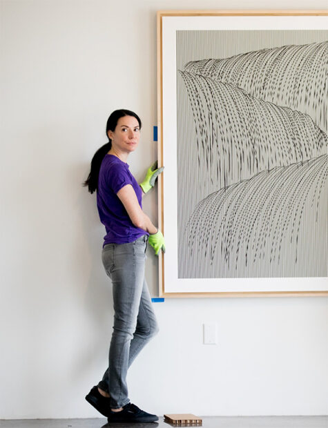 A photograph of Nancy Koen by Allison V. Smith. Nancy stands near a white gallery wall and holds the side of a framed work of art. She wears bright green gloves, a black t-shirt, and jeans.