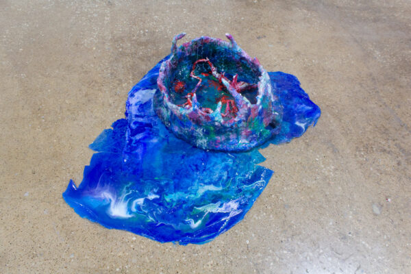 A blue sculpture of epoxy clay and resin formed into a bowl with clay forms sitting inside of it. A ragged sheet of dried resin sits underneath