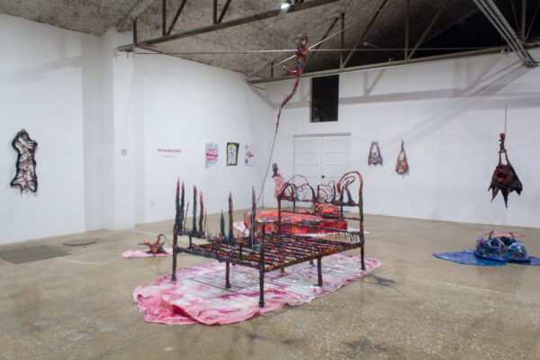 An interior view of a gallery with white walls. Sculptures made of epoxy clay sit on the wall, with a red bedframe covered in epoxy clay and sitting on a pink and white sheet of resin.