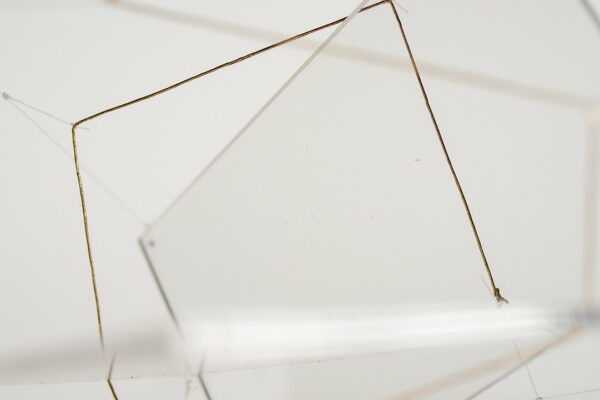 A close-up photograph of an installation by Ronald Llewellyn Jones. The image depicts lines of thread and a clear acrylic sheet suspended by thread.