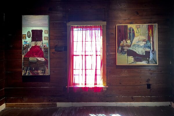 Sobre una pared de tablas, hay una ventana cubierta por una cortina semitransparente y roja. A sus costados hay un par de pinturas, ambas son retratos de personas mayores de tono de piel oscuro en camas hospitalarias. Los cuartos dentro de ambas pinturas tienen cortinas rojas.