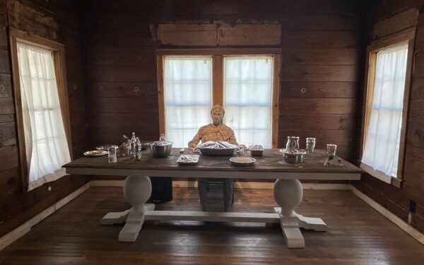Un cuarto con ventanas y cortinas blancas en tres de sus paredes. En el centro, una larga mesa de madera sobre la cual hay piezas de una vajilla metálica y pequeñas figuras cubiertas con aluminio. Frente a nosotros y al centro de la mesa está sentada una figura humana de papel maché.
