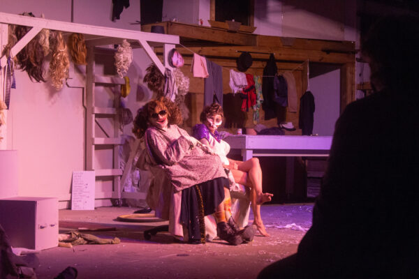 Two performers in wigs and makeup sit on chairs on a stage inside a warehouse amphitheater.