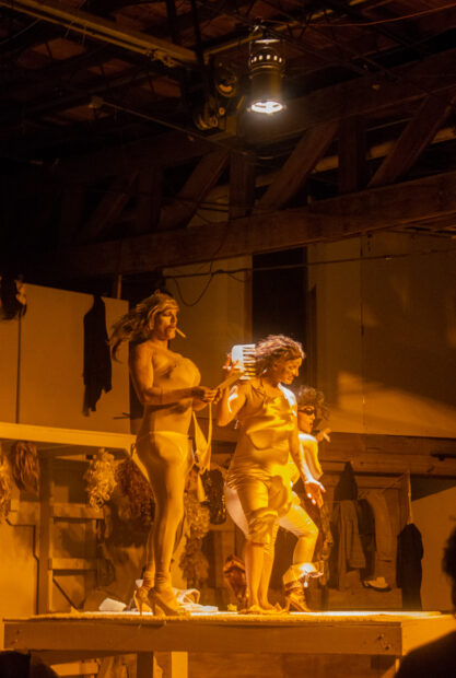 Three performers in gold spandex fabric and wigs stand on a stage underneath a bright spotlight.