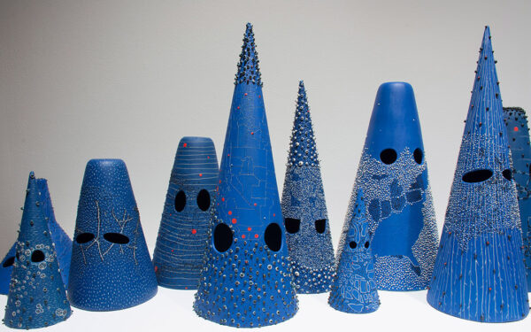 A photograph of an assortment of blue cones and cone-like objects set on a table. The objects, of varying sizes, each have a pair of circles or ovals removed from them so that they resemble a mask or something that would be worn on the head to cover the face.