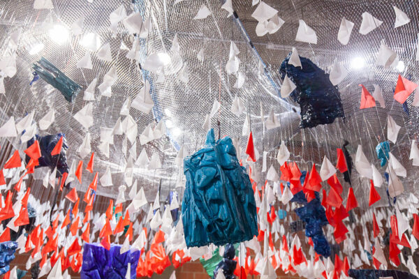 An photograph of a large installation work by Tammie Rubin. The work features a large fishing net hanging from the ceiling with small white and orange stake flags hanging upside-down from the net. In the fore is a bright blue abstract resin object.