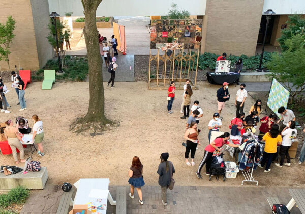 A photograph showing an installation by Tay Butler in the courtyard of the University of Houston's School of Art building. There is a crowd of people in the courtyard participating in various activities.