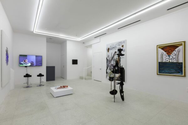 An installation image of "Texan: Part I," at VSF Texas. The image shows two canvases hanging on a white wall with two sculptures in the center of the room and a large screen mounted on one wall with headphones available for visitors.