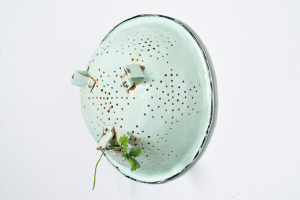 A sculpture by Tamara Johnson of a mint green colander with a fabricated piece of cilantro clinging to the bottom.