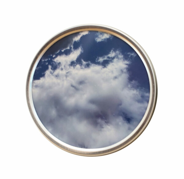 A close-up image of a small circular photograph featuring white clouds set against a blue sky. The photograph is housed in a round silver tin. Artwork by Mery Godigna Collet.