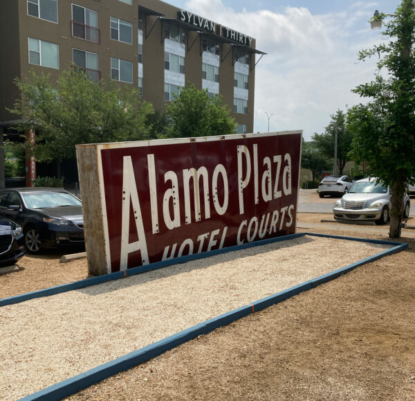 A red and white metal sign that reads "Alamo Plaza Hotel Courts", which is installed in the parking lot of the Sylvan Thirty Apartment building