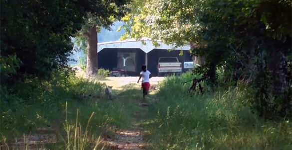 A young Black person is running away from the viewer, through a dirt pathway lined with mature trees. They are running towards a double-wide garage with an an antique truck parked in the right bay of the garage