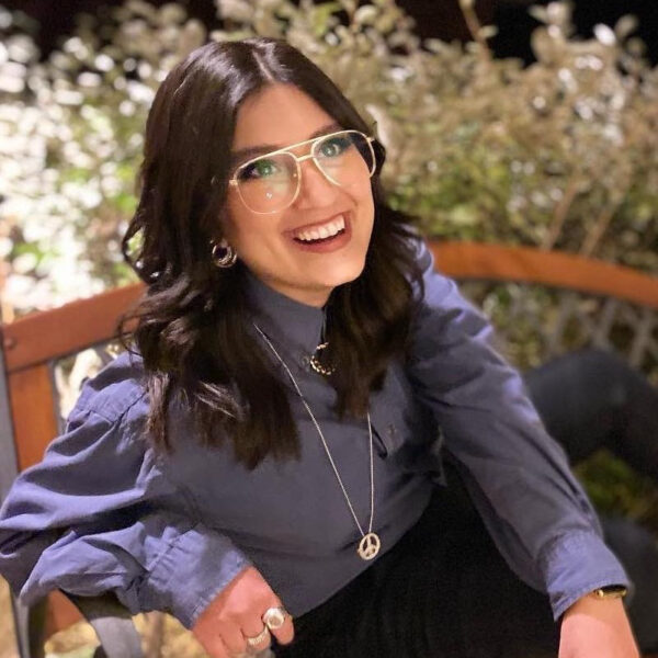 A photograph of Asaiah Puente. She wears a long sleeve button up shirt, wire frame glasses, and a long necklace. She looks up and to the right with a wide smile.