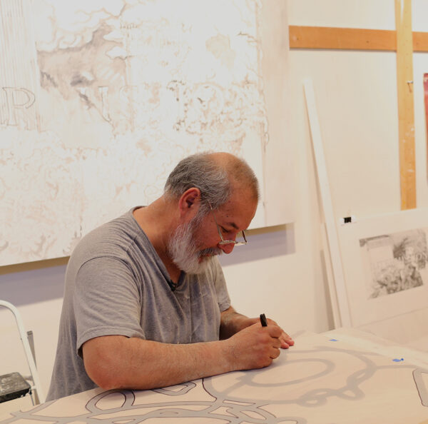 A photograph of artist Benito Huerta working in the studio. He sits at a table and traces an drawing on thin paper.