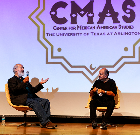 A photograph of Benito Huerta and Cheech Marin sitting in chairs on a stage. The text projected on the screen behind them reads, "CMAS Center for Mexican American Studies. The University of Texas at Arlington."