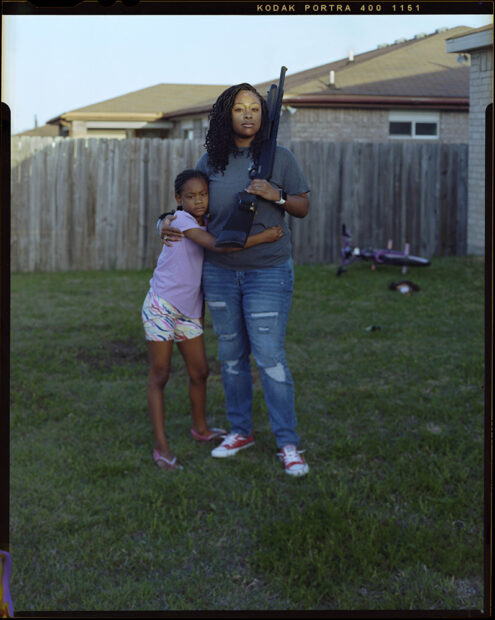 A photograph of a Black mother with her arm around her pre-teen daughter. They stand in their backyard and the mother olds a large gun in her other hand. Photograph by Christian K. Lee.