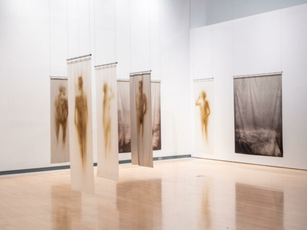 An installation view of work by Oscar Muñoz. The image shows an arrangement of shower curtains, which have been painted with blurred figures, hanging from the ceiling.