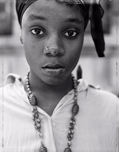 A black and white photography by Dawoud Bey of a young Black girl. She looks straight into the camera and wear a small nosepin jewelry piece that looks like a tiny knife. 